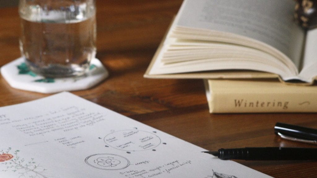 Close up of a wood desk with a book open, a glass of water and a notebook with handwritten notes and drawings.